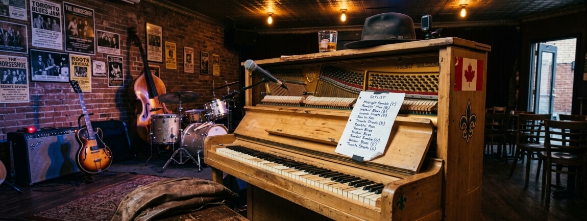 Upright piano with handwritten setlist, guitar, upright bass, and drums on a Toronto blues bar stage with vintage concert posters on exposed brick